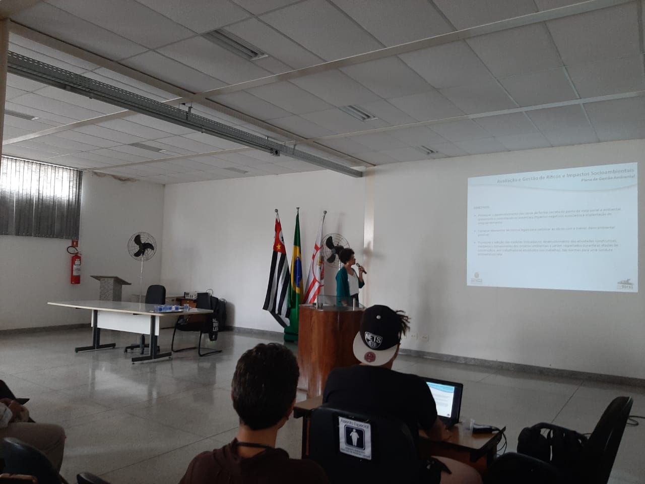 Imagem de Andrea Franklin apresentando o projeto em uma projeção na parede, com um microfone, em uma tribuna. Pessoas ao primeiro plano olhando para a projeção
